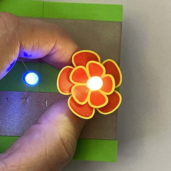 A bright light in the middle of a red and yellow printed flower. This is being held by a hand over 2 silver lines of tape on a green foam block