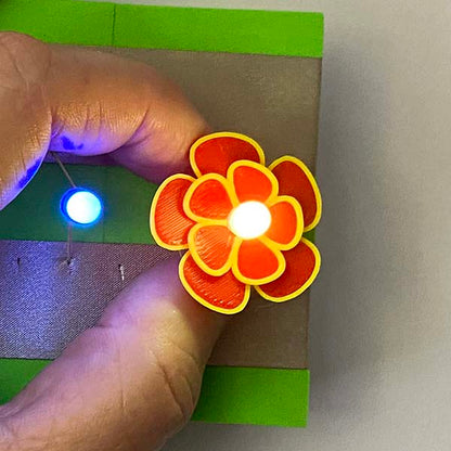 A bright light in the middle of a red and yellow printed flower. This is being held by a hand over 2 silver lines of tape on a green foam block