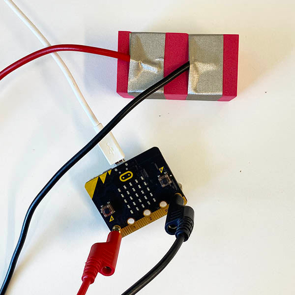 A close up of a red MicroBit connector TapeBlock. Below this is a Micro:bit  with a red banana clip attached to the pin 0 and Black banana plug attached to ground. The Micro:bit is also connected to a white Micro USB lead