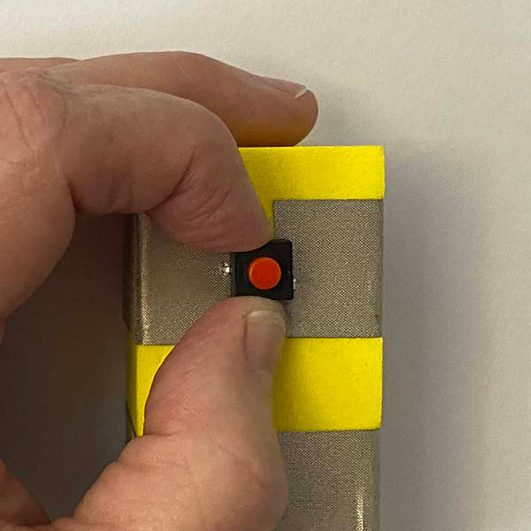 Close up of fingers holding a black and red button between two pieces of silver tape which are around a yellow foam block