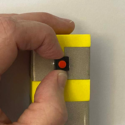 Close up of fingers holding a black and red button between two pieces of silver tape which are around a yellow foam block