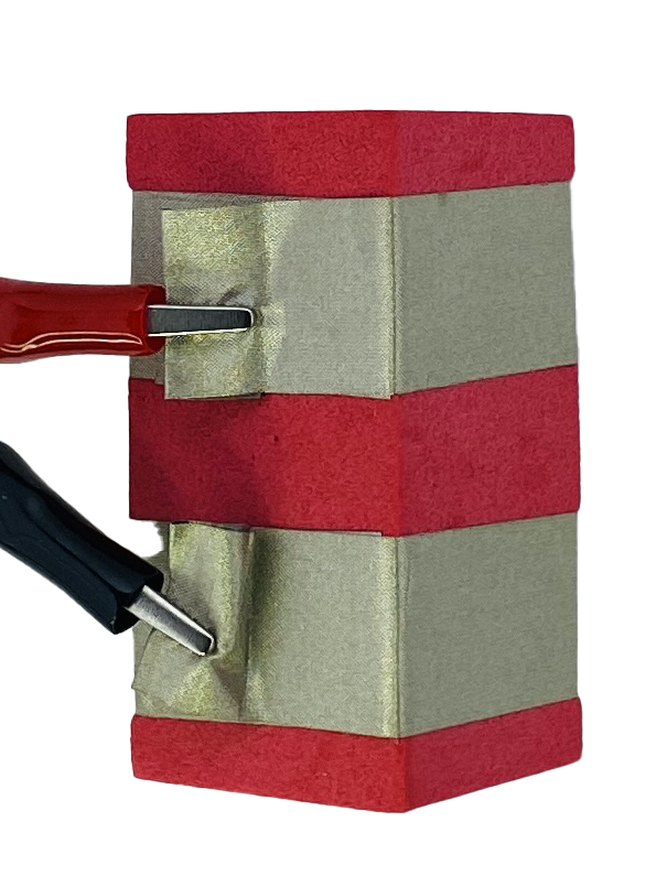 A red foam block with two parallel rows of conductive tape wrapped around it. Each of the rows has a tab of extra tape that has a red or black alligator clip attached to it. 