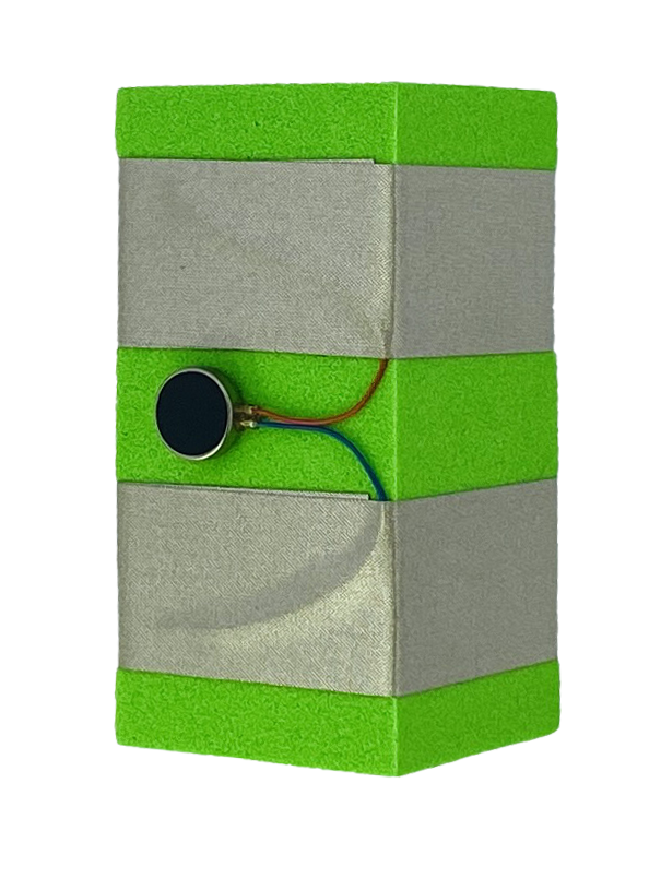 A green foam block with two parallel rows of conductive tape wrapped around it. There is a vibration motor which has a blue and red wire connected to the two pieces of tape. 