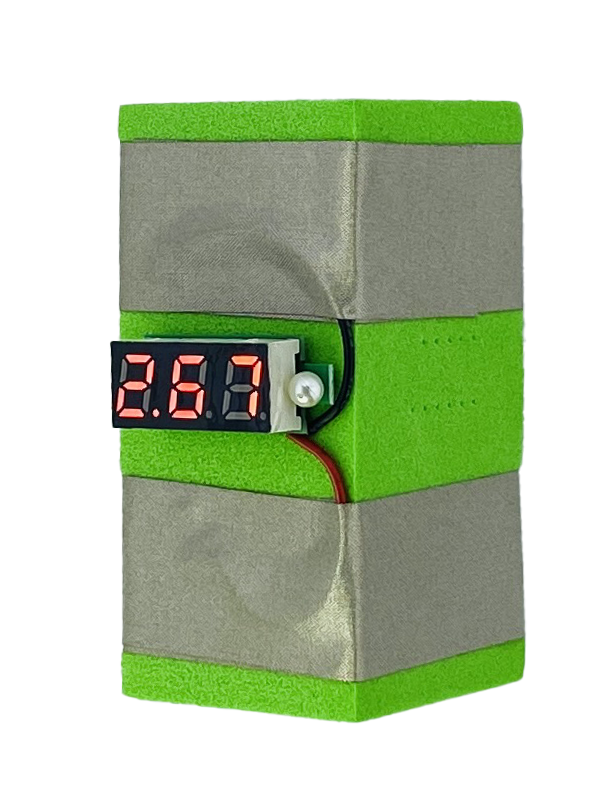 A green foam block with two parallel rows of conductive tape wrapped around it. There is a voltage display which is black and displays the red numbers 2.67. It has a blue and red wire connected to the two pieces of tape. 