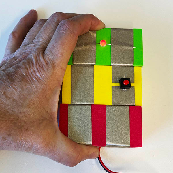 A close up of a hand holding 3 TapeBlocks together. Green with red light, yellow with a button and red attached to the USB cable TapeBlock