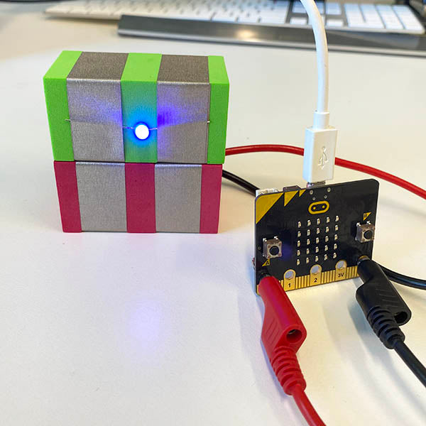 A close up a Green light TapeBlock sitting on a red TapeBlock MicroBit connector TapeBlock. In front of this is a Microbit with a red banana clip attached to the pin 0 and Black banana plug attached to ground.