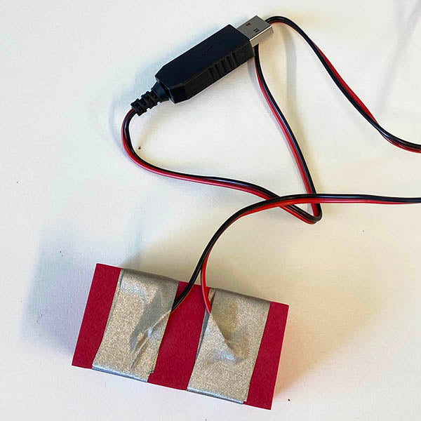 A red foam TapeBlock with a red an black wire clearly placed under the 2 parallel pieces of tape. The wires are from a chunky USB power connector