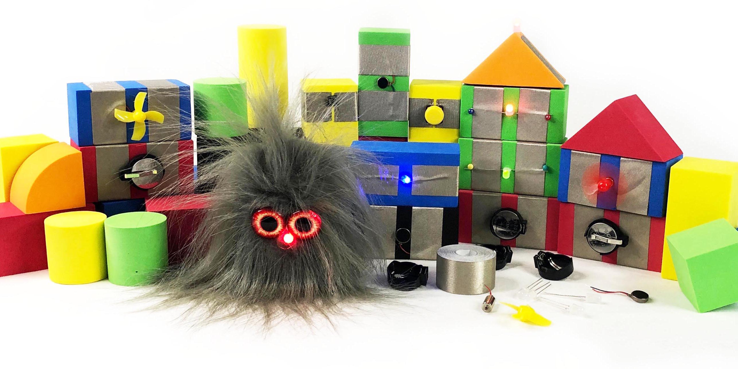 Left to right foam block of different shapes, a fan TapeBlock, a furry TapeBlock character with a red light up nose, a Viabration TapeBlock, a Switch TapeBlock, A fan TapeBlock, in front components including LEDs and battery carriers. Conductive tape