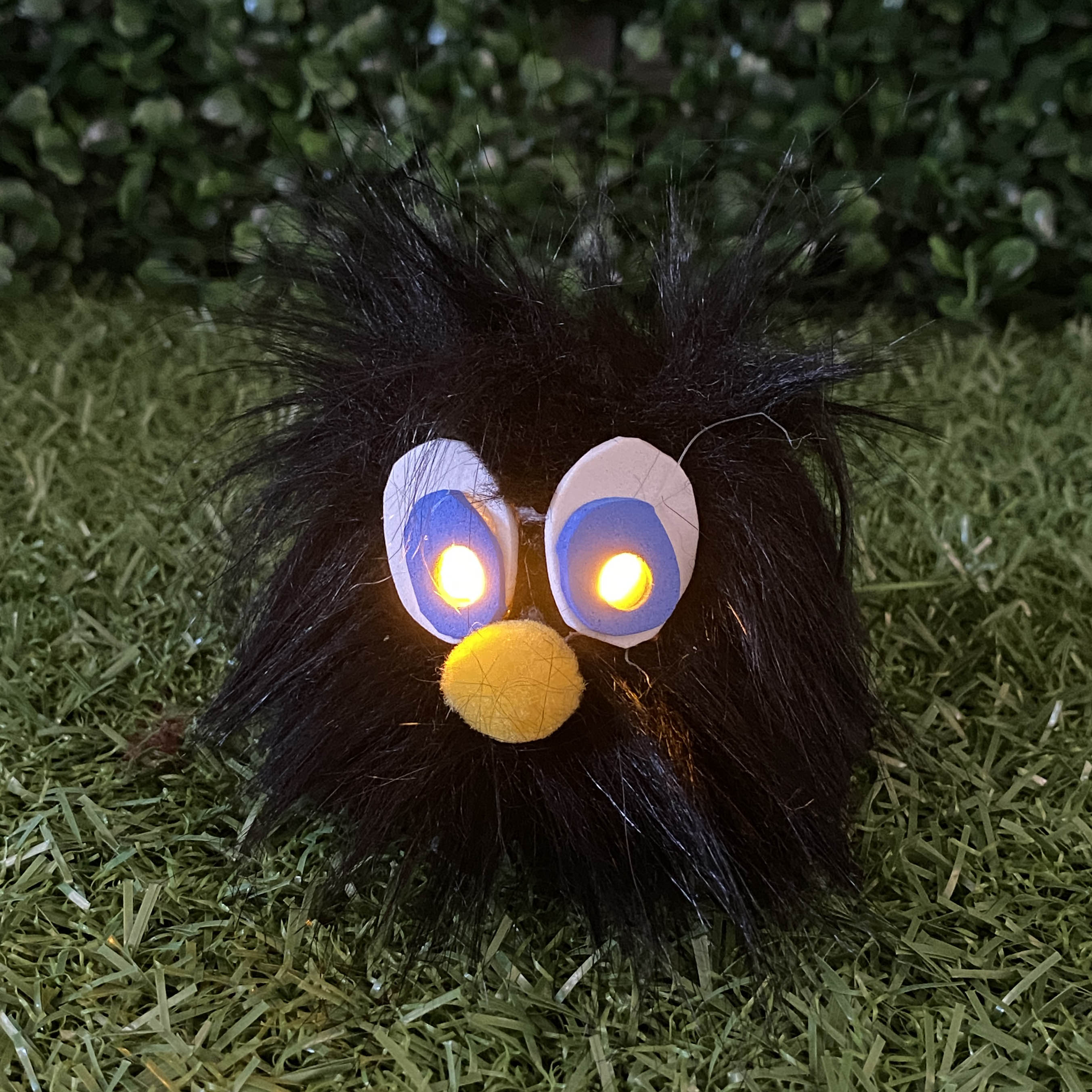 Black fur TapeBlock with yellow  light up eyes and yellow pom pom nose, sitting on the grass