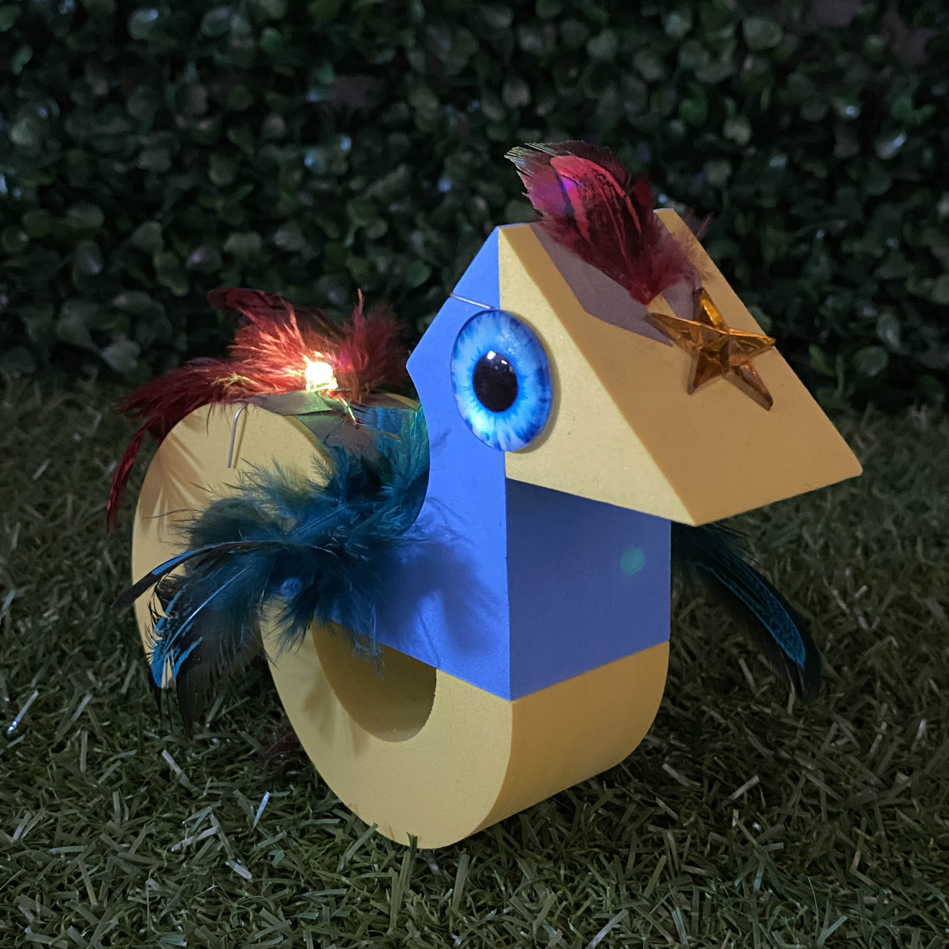 Bird shaped TapeBlock with blue eyes and yellow light up tail, it is sitting on the grass