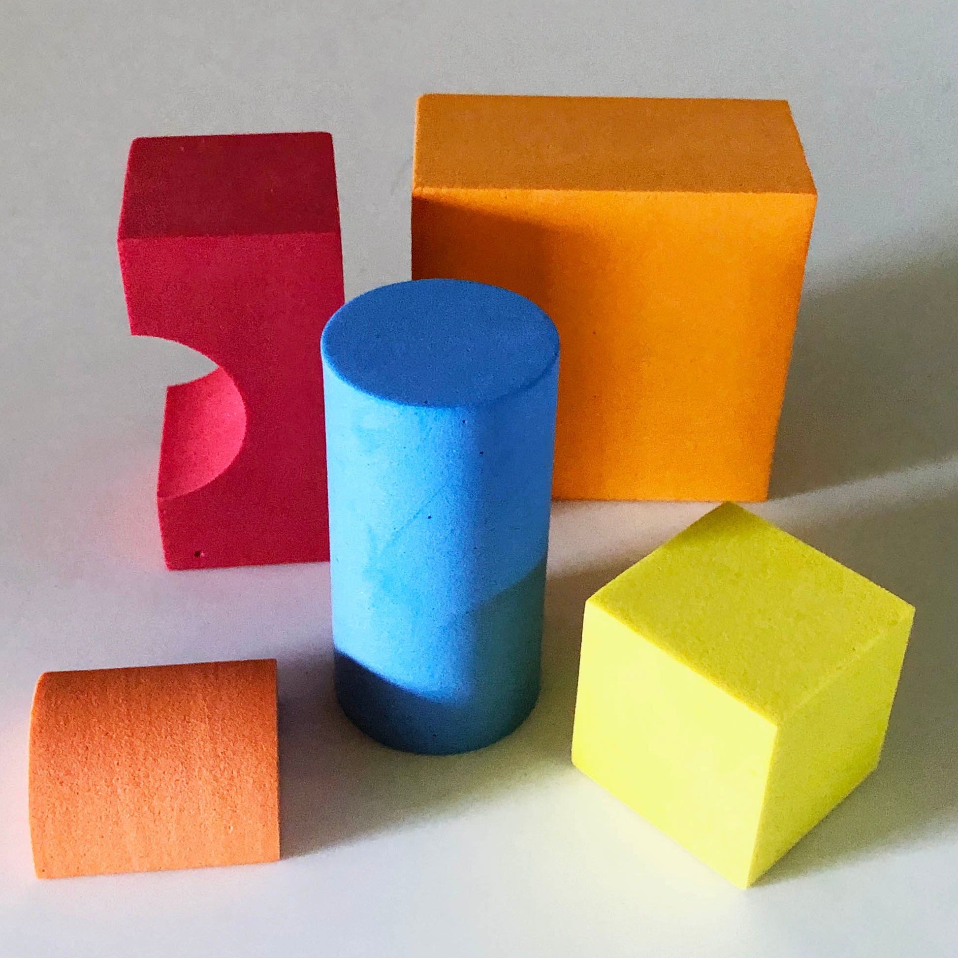 Odd shaped and colored foam blocks including: Red arch, orange shape with a square face and rectangle top, blue cylinder, orange small half cylinder and yellow square cube